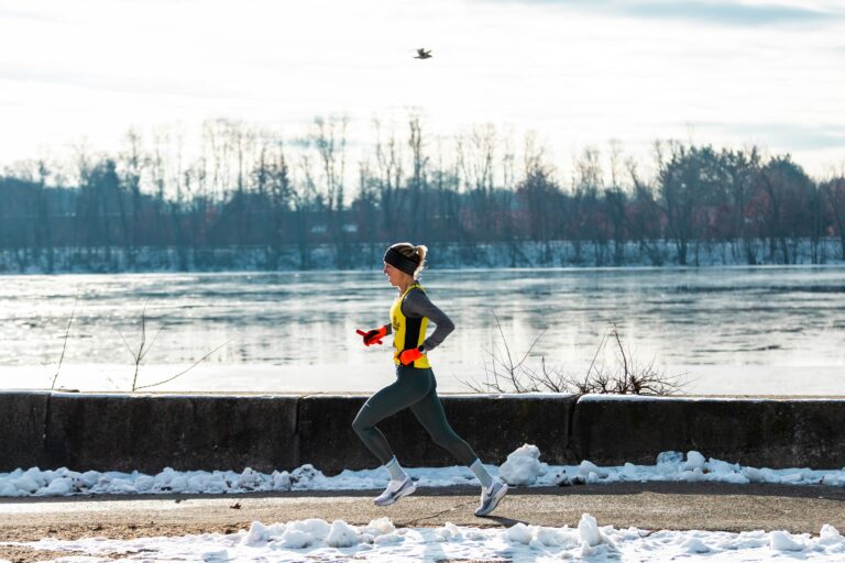 female runner in winter gear with gloves and reflective vest running along a snowy riverbank